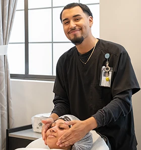 Male cosmetology student smiles as he massages the face of a female patron.